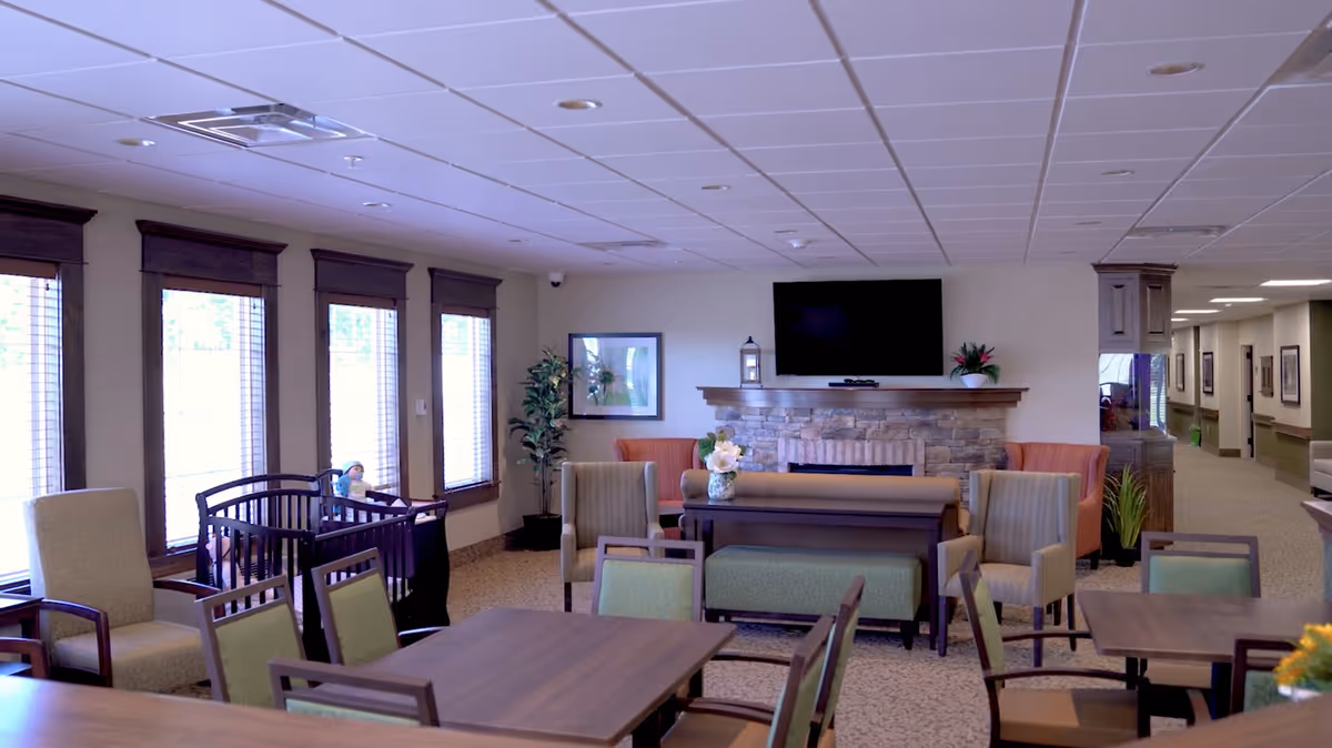 Bright communal dining and lounge area with tables and chairs, a fireplace, TV, and large windows.