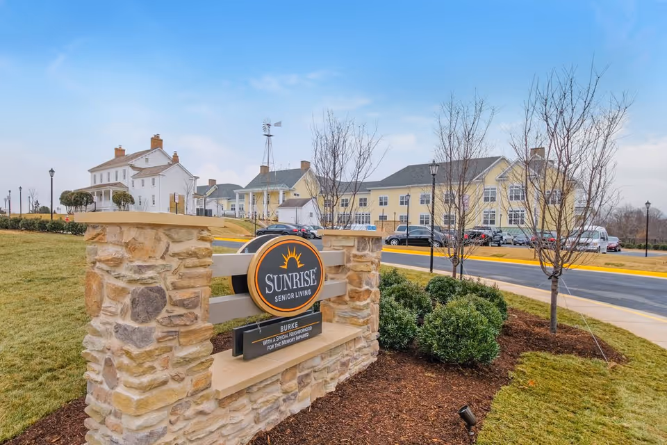 Stone monument sign reading 'Sunrise Senior Living' in front of a landscaped driveway and the facility's yellow and white buildings.