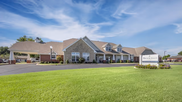 Exterior view of Brookdale Chickasha senior living facility with a large green lawn in the foreground, a clear blue sky with some clouds, and a building featuring a covered entrance and multiple windows.