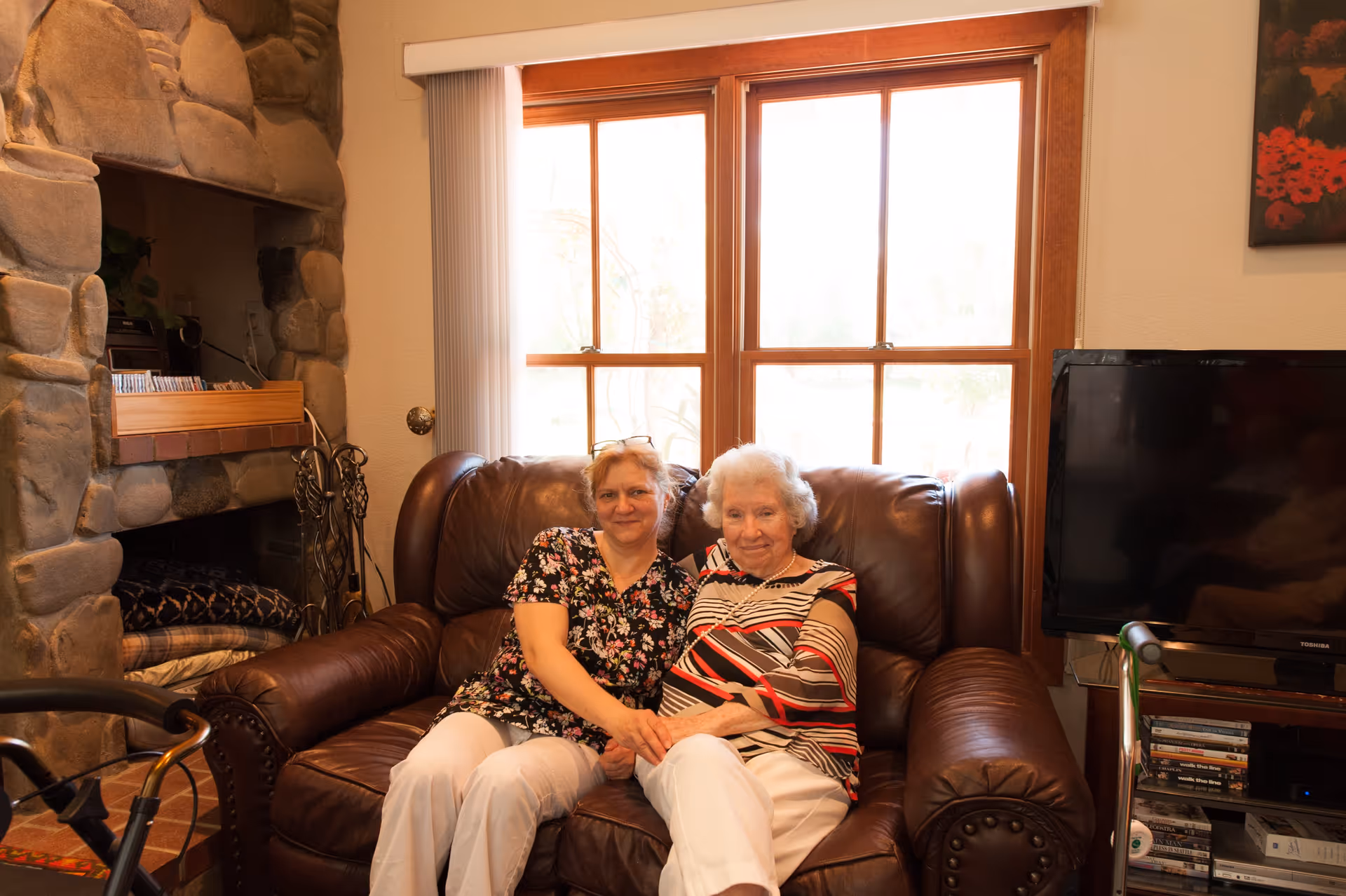 Two women sitting closely together on a brown leather couch in a cozy living room. Behind them is a large window with wooden frames letting in natural light. To the left is a stone fireplace with some items on the mantel, and to the right is a flat-screen TV on a stand with shelves holding DVDs and other items.
