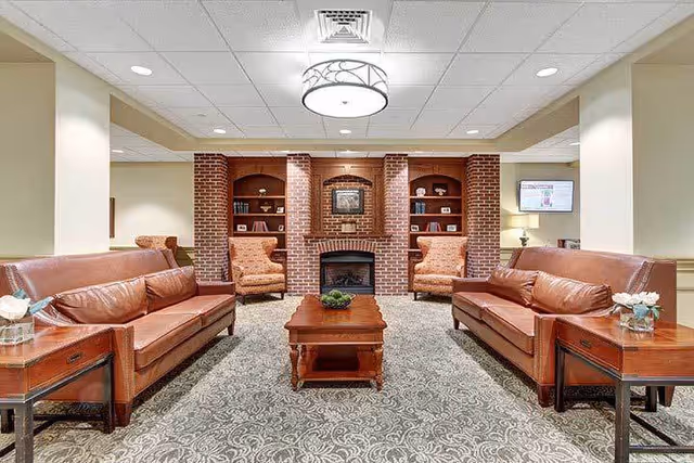 A cozy living room area with two brown leather sofas facing each other, a wooden coffee table in the center, and two upholstered armchairs near a brick fireplace with built-in shelves on either side. The room has a patterned carpet, soft lighting, and neutral-colored walls.