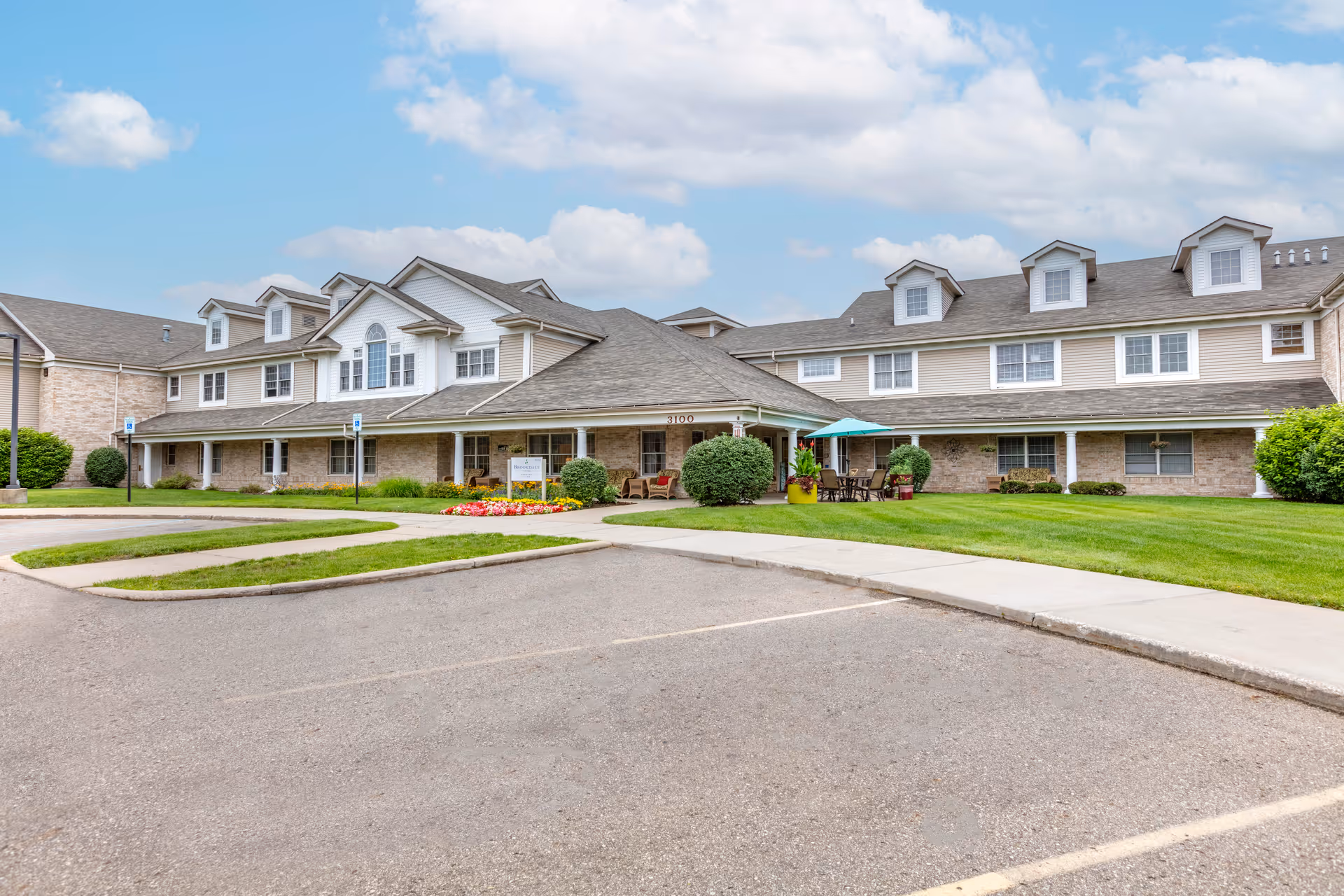 Exterior view of a two-story senior living facility building with a large parking area in front, well-maintained green lawn, flower beds, and outdoor seating with an umbrella. The building has multiple windows and dormer windows on the roof under a partly cloudy sky.
