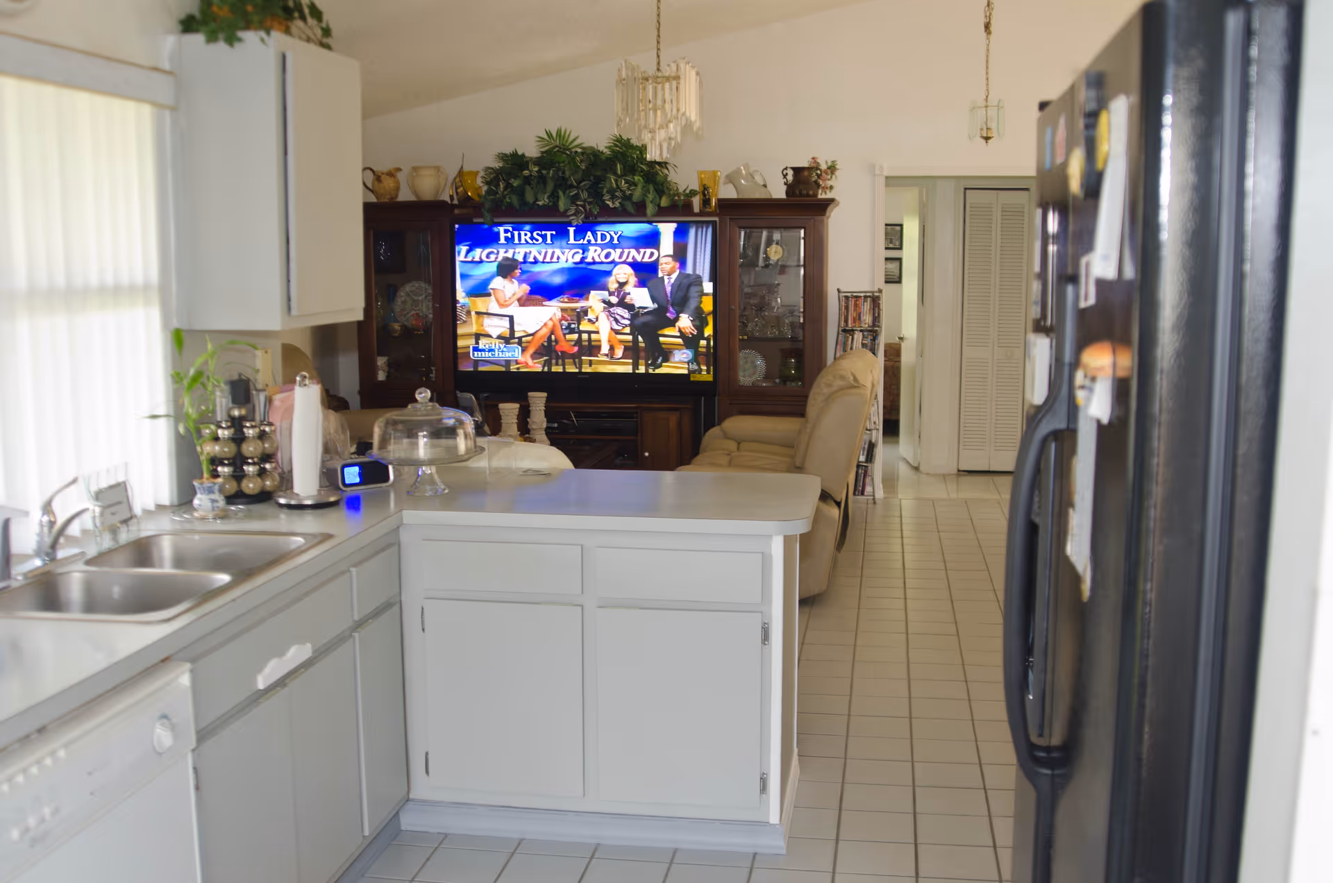 View of a kitchen with white cabinets, a double sink, and a dishwasher on the left. A black refrigerator is on the right. Beyond the kitchen is a living room area with beige recliner chairs, a wooden cabinet with glass doors, and a television showing a talk show titled 'First Lady Lightning Round'. The floor is tiled in white, and there are plants and decorative items on top of the cabinets.