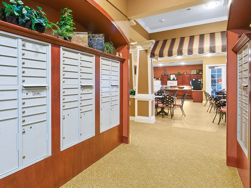 Interior common area showing wall-mounted resident mailboxes opening into a communal dining/lounge area with tables, chairs, and a kitchenette.