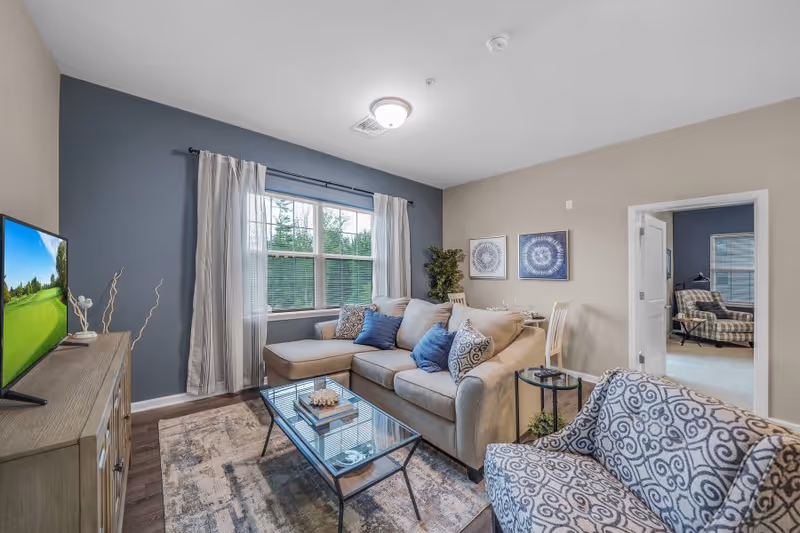 Bright living room with a beige sectional sofa, patterned armchair, glass coffee table, TV and a window with curtains.