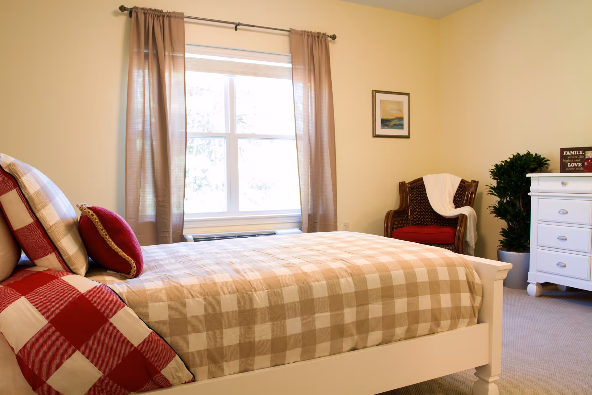 A cozy bedroom with a white wooden bed featuring beige and red checkered bedding and pillows. There is a large window with beige curtains letting in natural light. A wicker chair with a red cushion and a white throw blanket is placed in the corner next to a green potted plant. A white dresser with drawers is also visible, along with a framed picture on the wall.