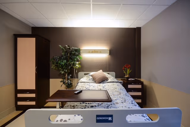 A neatly made single hospital-style bed in a private room with bedside tables, a wardrobe, a potted plant, and an over-bed table.