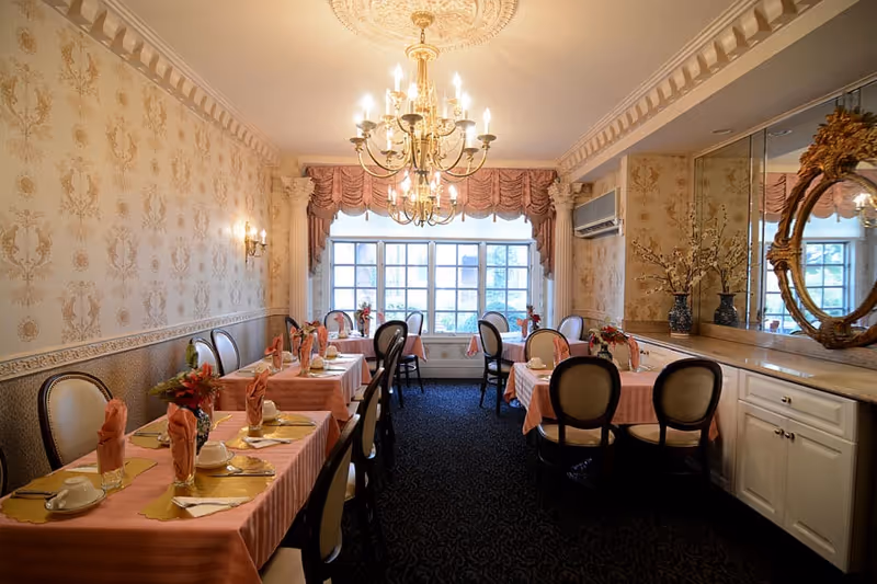 Elegant dining room with several tables covered in pink tablecloths, each set with folded napkins, cups, and plates. The room features ornate wallpaper, a large chandelier, a window with draped curtains, and a mirrored wall with decorative vases and a gold-framed mirror.