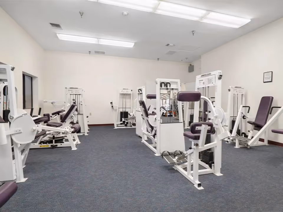 Carpeted fitness room with white strength-training machines and purple-upholstered seats under fluorescent lights.