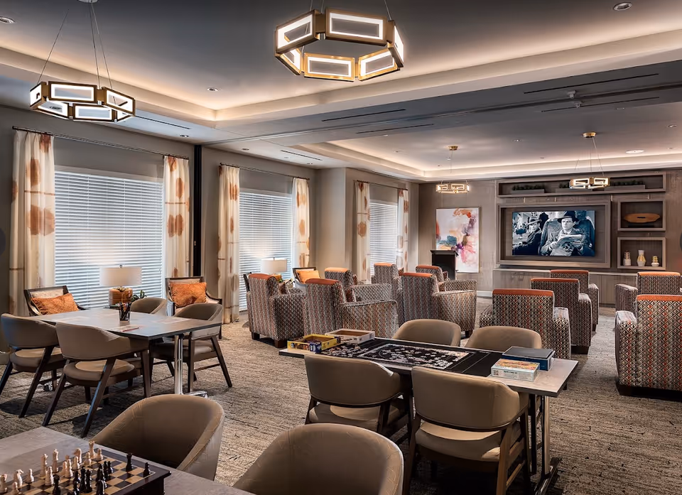 A spacious and well-lit common area in a senior living facility with multiple seating arrangements including patterned armchairs facing a large wall-mounted TV showing a black and white movie. Several tables with chairs are set up for games such as chess and puzzles. The room features modern light fixtures, large windows with blinds and patterned curtains, and decorative artwork on the walls.