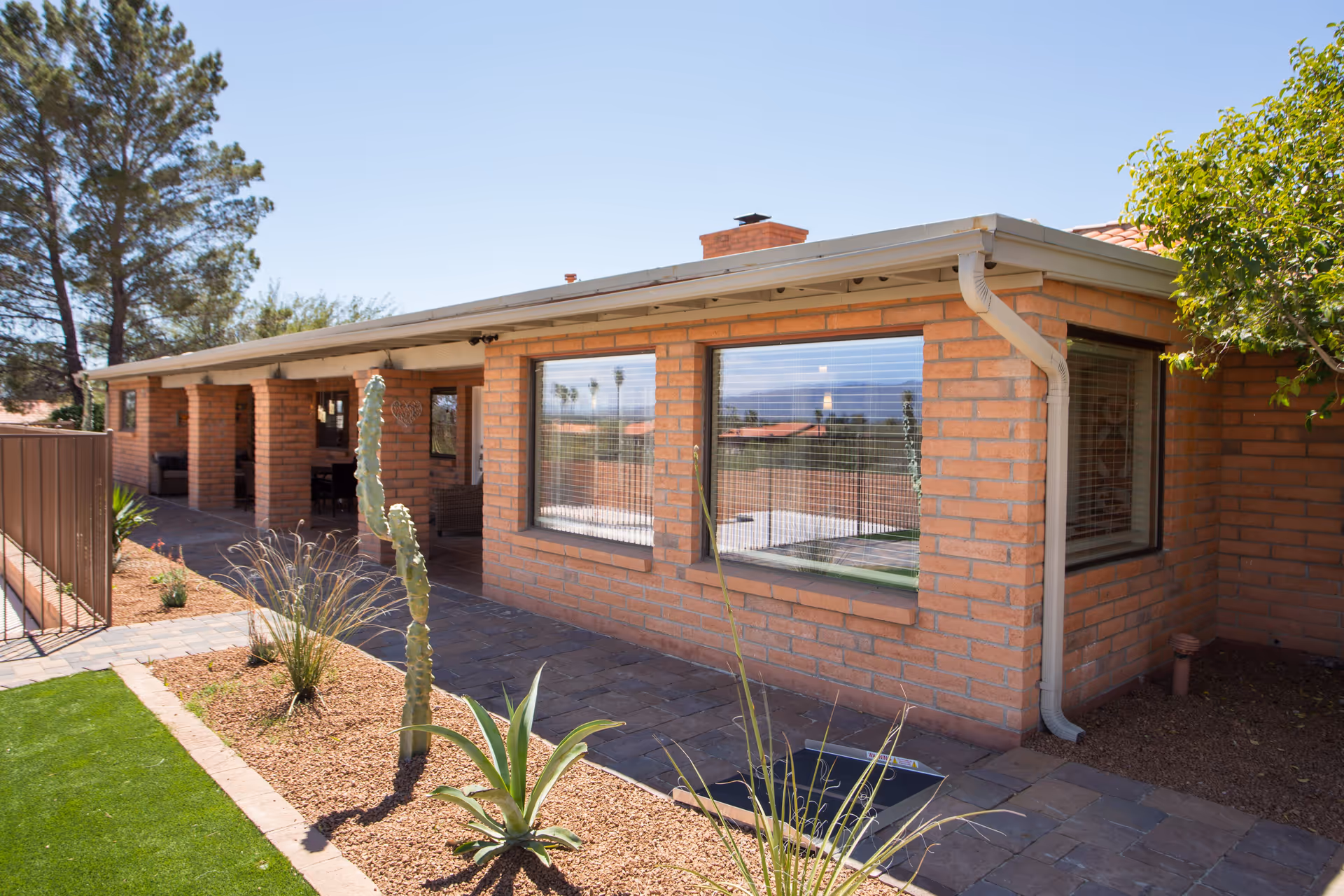 Exterior view of a single-story brick building with large windows and a covered patio area. The landscaping includes desert plants such as cacti and agave, with a paved walkway and a green lawn area under a clear blue sky.