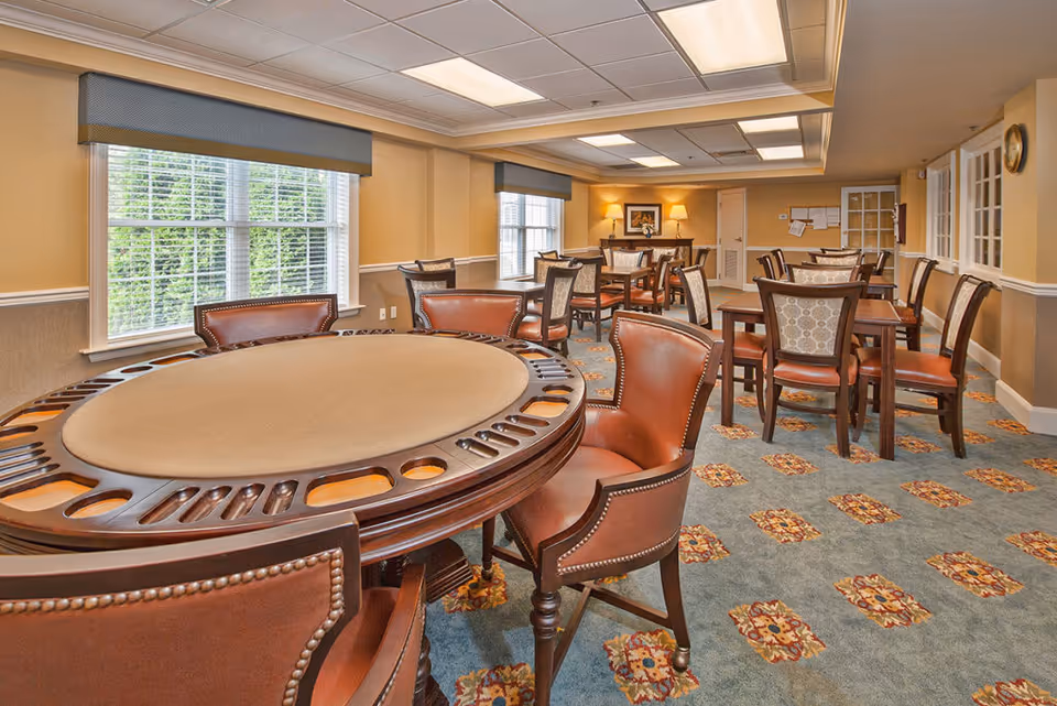 A well-lit room with multiple tables and chairs arranged for group seating. The foreground features a round table with leather chairs, likely designed for card games or social activities. The room has large windows with blinds, beige walls, patterned carpet, and ceiling lights providing bright illumination.