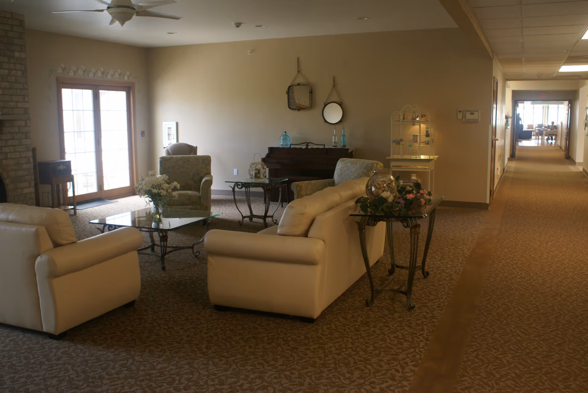 A cozy living room area with beige leather sofas and patterned armchairs arranged around a glass coffee table. There is a piano against the wall with decorative items on top, two round mirrors hanging above it, and a birdcage-style decorative piece nearby. The room has carpeted flooring and a ceiling fan, with a hallway extending to the right leading to another room with tables and chairs.