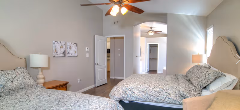 A senior living facility bedroom with two beds featuring patterned bedding, a wooden nightstand with a lamp, ceiling fans with lights, and neutral-colored walls decorated with floral artwork. The room has an open doorway leading to another area with additional doors visible.