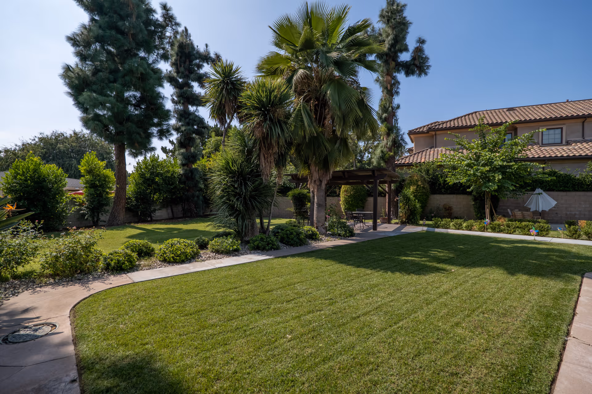 A well-maintained outdoor garden area with green grass, various trees including palm trees and pine trees, shrubs, and a paved walkway. There is a wooden pergola with seating underneath and a two-story building with a tiled roof in the background.