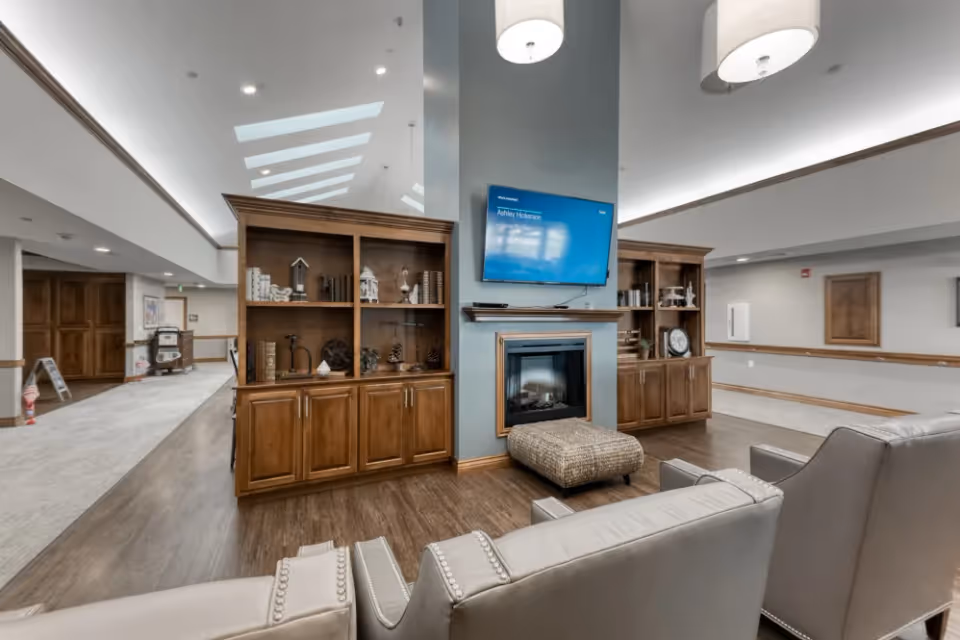 A cozy common area in Addington Place of Sparta featuring a wall-mounted flat-screen TV above a fireplace, flanked by two wooden bookshelves with decorative items. In front of the fireplace is a cushioned ottoman, and there are several upholstered chairs arranged facing the TV. The space has wood flooring and a hallway with carpet and wooden doors in the background.