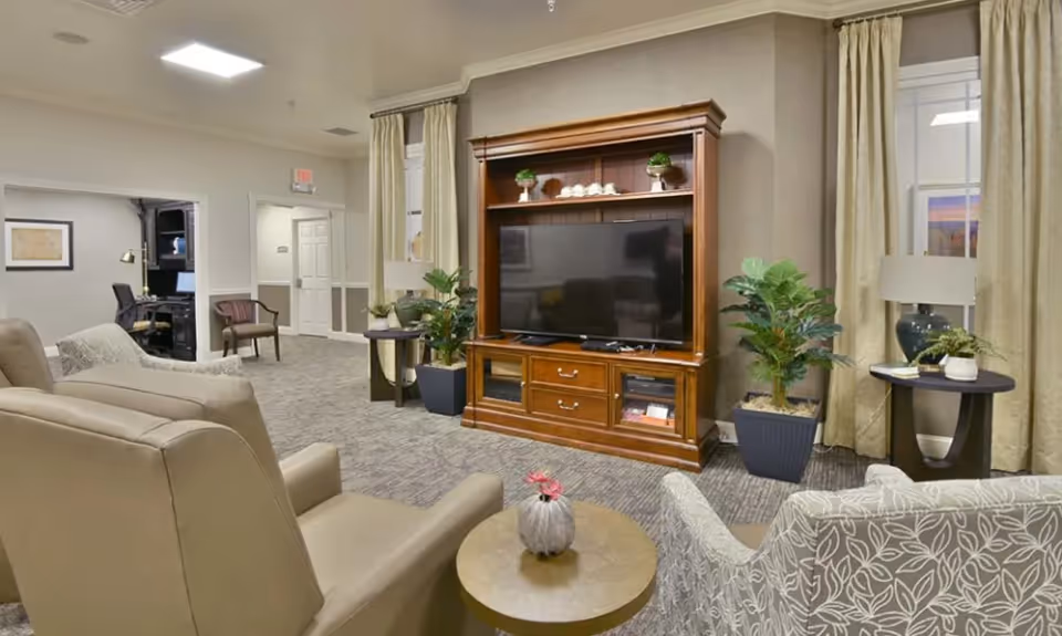 A cozy senior living facility common area with beige armchairs and a patterned armchair arranged around a small round table with a decorative vase. A large wooden entertainment center with a flat-screen TV and decorative plants is against the wall. The room has carpeted flooring, beige walls, and windows with light-colored curtains. In the background, there is an office area with a desk and chair.