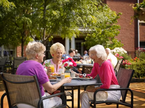 Three elderly women sit at a sunlit outdoor patio table, enjoying food and drinks and laughing.