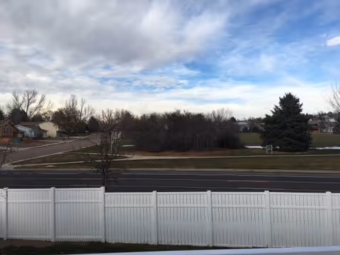 Residential neighborhood view with a white fence, road, trees, and cloudy sky.