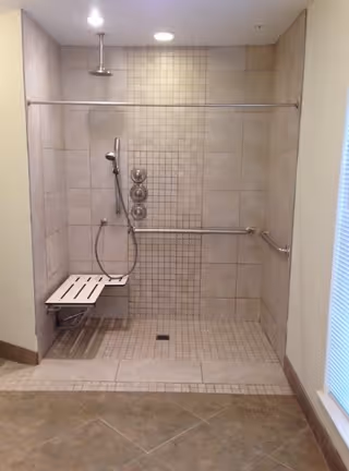 A tiled walk-in shower with a fold-down bench, handheld showerhead, overhead shower, and grab bars on the walls. The shower area has a tiled floor with a drain in the center and is well-lit with ceiling lights.