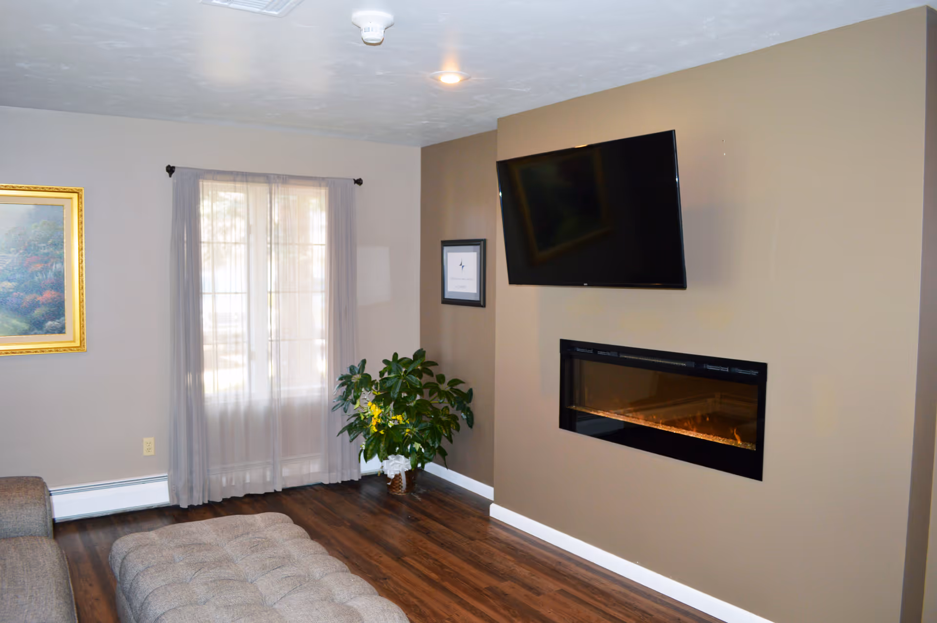 A cozy living room with a wall-mounted flat-screen TV above a modern electric fireplace. There is a large window with sheer curtains, a green potted plant in the corner, a framed picture on the wall, and a cushioned ottoman on the wooden floor.