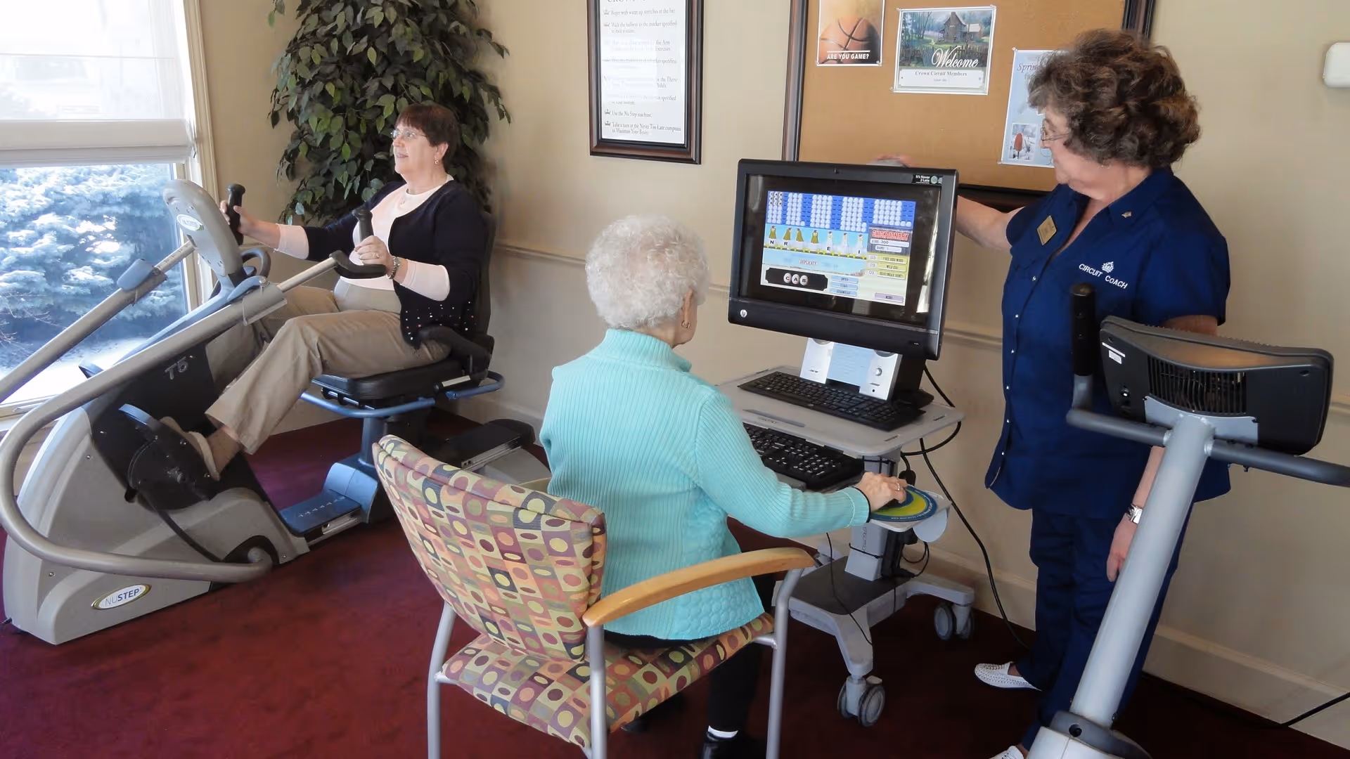 An elderly woman in a turquoise sweater sits in a patterned chair using a computer with a large screen, while another elderly woman exercises on a recumbent bike near a window. A staff member in a navy blue uniform labeled 'Circuit Coach' stands next to a treadmill, pointing at the computer screen in a room with beige walls and red carpet.