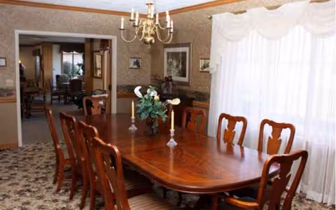 Senior living facility dining room with a long wooden table, chairs, chandelier, and centerpiece.