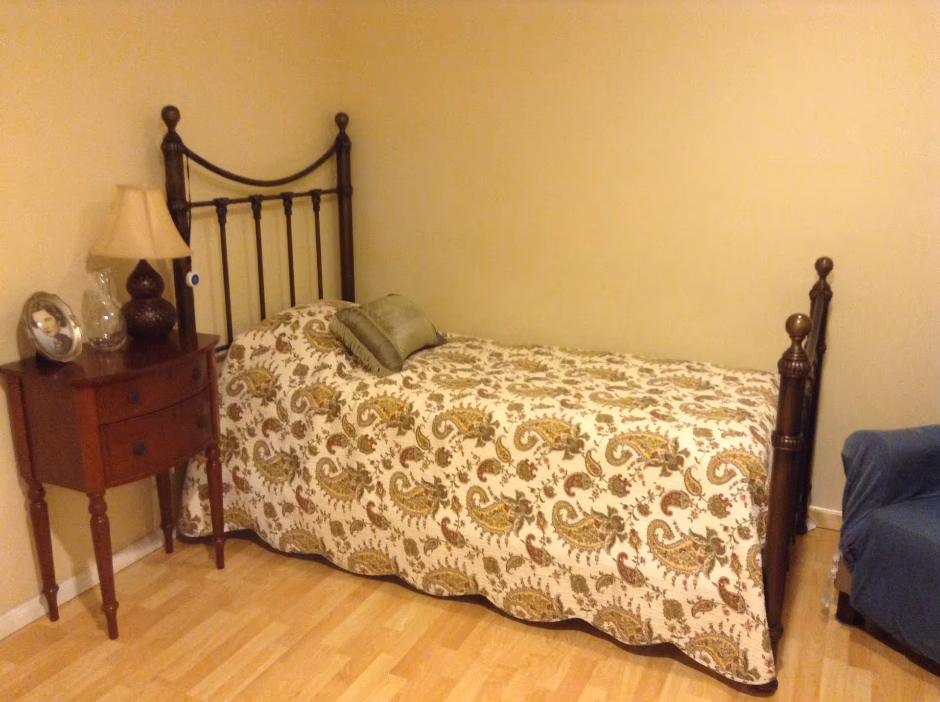 A small bedroom with a single bed featuring a dark metal headboard and footboard. The bed is covered with a patterned quilt and has two green pillows. Next to the bed is a wooden nightstand with two drawers, holding a table lamp, a glass vase, and a framed photo. The room has light-colored walls and a wooden floor. A blue upholstered chair is partially visible on the right side.