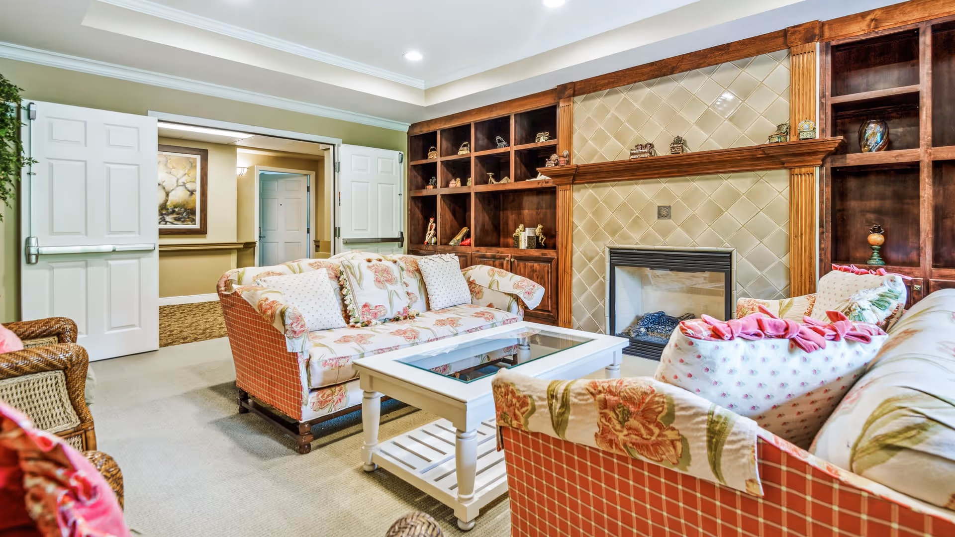 A cozy living room with floral patterned sofas and a white coffee table in the center. The room features a fireplace with a tiled surround and wooden built-in shelves on either side, decorated with small ornaments. Double white doors open to a hallway with framed artwork on the wall.