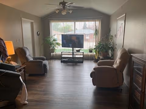 Spacious living room with three recliners facing a TV in front of a large window and potted plants.