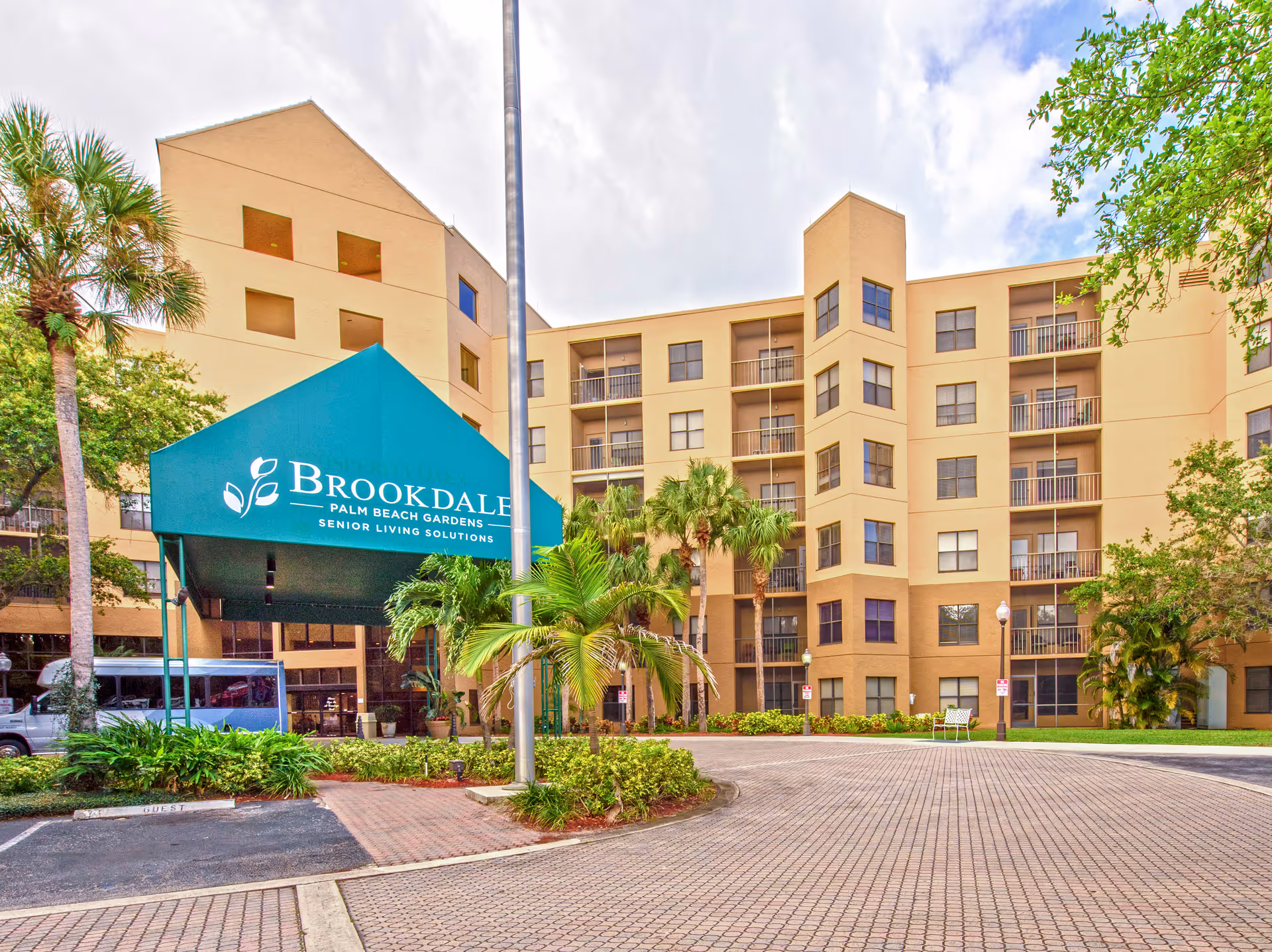 Front entrance of the Brookdale Palm Beach Gardens senior living building with a green canopy, palm trees, and a multi-story beige facade.