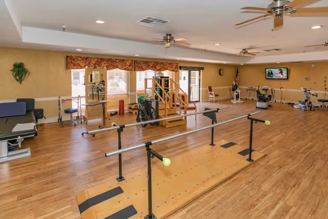 A spacious rehabilitation room with wooden floors, parallel bars for walking practice, exercise equipment including a wheelchair, stationary bikes, and a small staircase for physical therapy. The room has ceiling fans, large windows with blinds, and a wall-mounted TV.