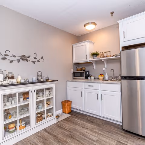 A small kitchen area with white cabinets, a stainless steel refrigerator, a microwave on the countertop, a sink, and various kitchen items including jars, a fruit bowl, and a soap dispenser. There is a white cabinet with glass doors displaying dishes and cups, a decorative wall piece above it, and a small trash bin on the floor. The flooring is wood-style and the walls are painted light beige.
