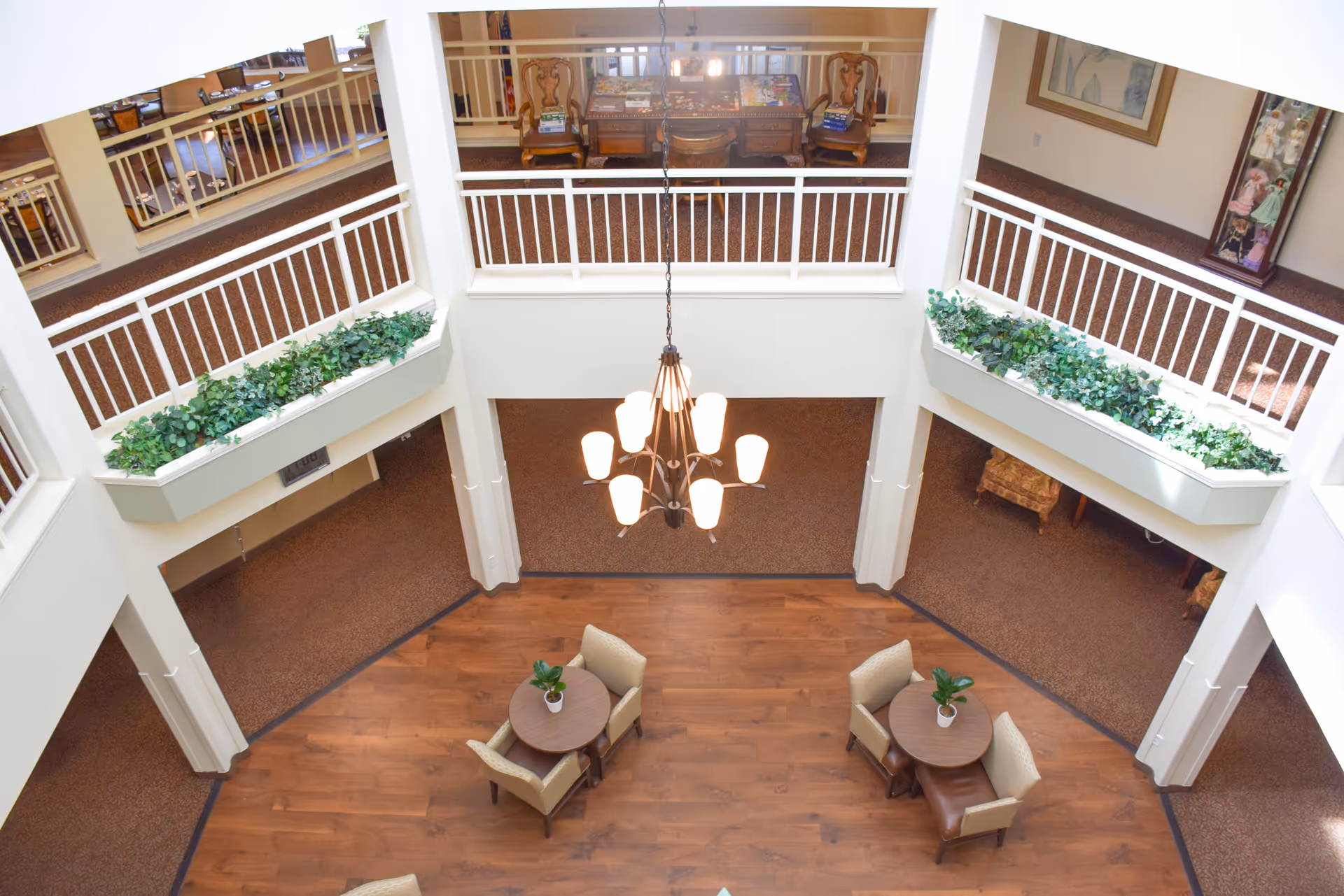 View from above of a two-story interior space in a senior living facility with a wooden floor area featuring two round tables, each with two chairs and a small plant. The upper level has white railings and planter boxes with green plants. A chandelier with multiple lights hangs in the center.