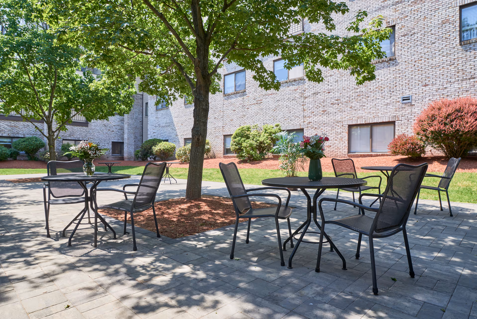 Outdoor patio area with metal tables and chairs arranged on a paved surface under the shade of trees. The background shows a brick building with windows and landscaped bushes and grass.