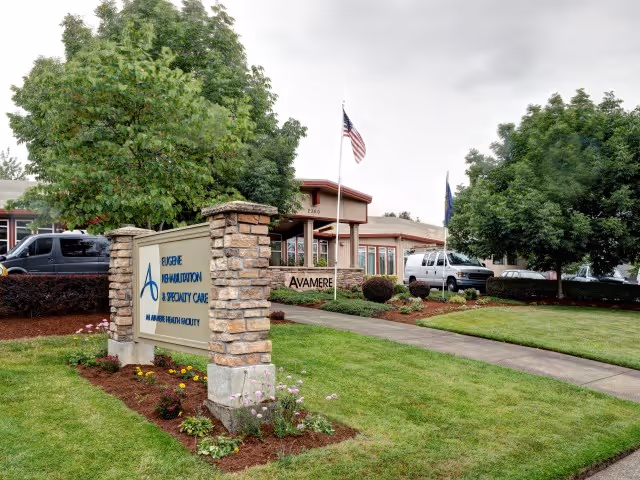 Exterior view of Avamere Rehabilitation of Eugene facility with a stone and wood sign in the foreground displaying the facility name and services. The building is surrounded by green grass, landscaped bushes, and trees. An American flag is flying near the entrance, and several vehicles are parked nearby under a cloudy sky.