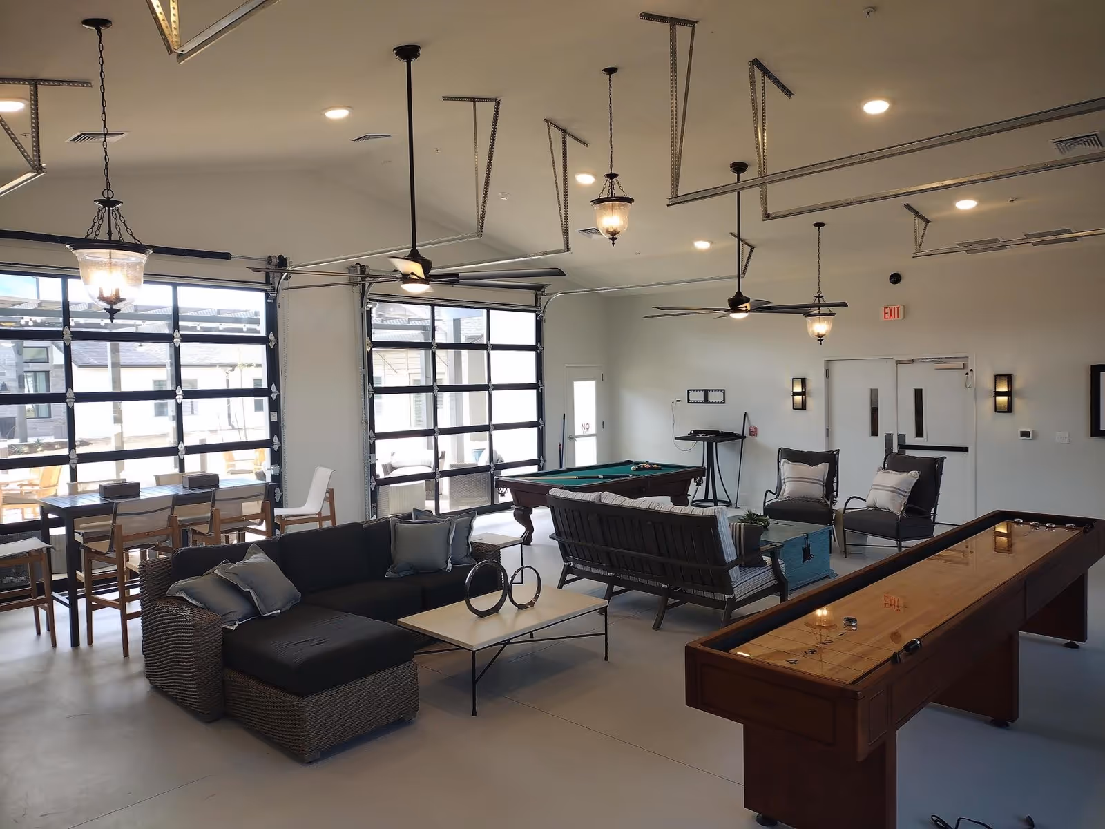 A spacious and well-lit common area with modern furniture including a sectional sofa, armchairs, and a coffee table. The room features a shuffleboard table, a pool table, and a dining table with chairs near large glass garage-style doors that open to an outdoor patio area. Ceiling fans and hanging light fixtures provide lighting and ventilation.