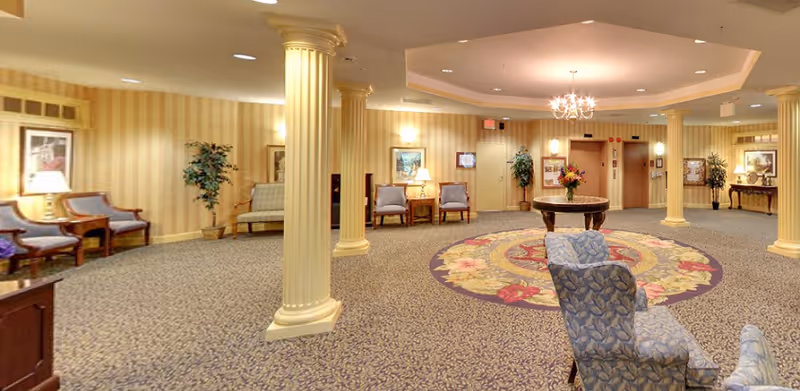 Spacious senior living facility common area with patterned carpet, decorative columns, multiple seating arrangements including armchairs and sofas, a round table with a floral centerpiece, warm lighting from wall sconces and a chandelier, and framed artwork on the walls.