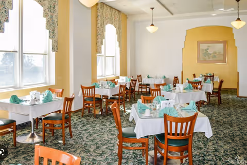 A bright dining room with multiple tables covered in white tablecloths and set with green folded napkins, cups, and saucers. Wooden chairs with green cushions surround the tables. Large windows with floral valances let in natural light, and the room features patterned green carpet and white walls with a yellow accent wall and framed artwork.