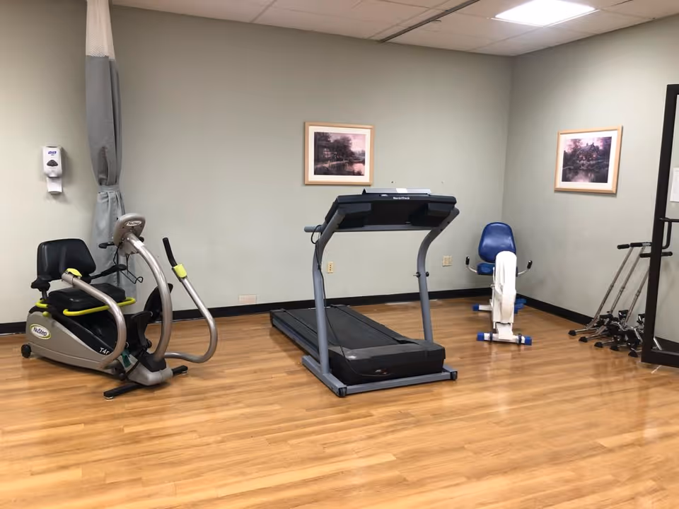 A small exercise room with wooden flooring containing a recumbent exercise bike, a treadmill, and a seated arm exercise machine. There are two framed landscape pictures on the walls and a hand sanitizer dispenser mounted on the left wall.