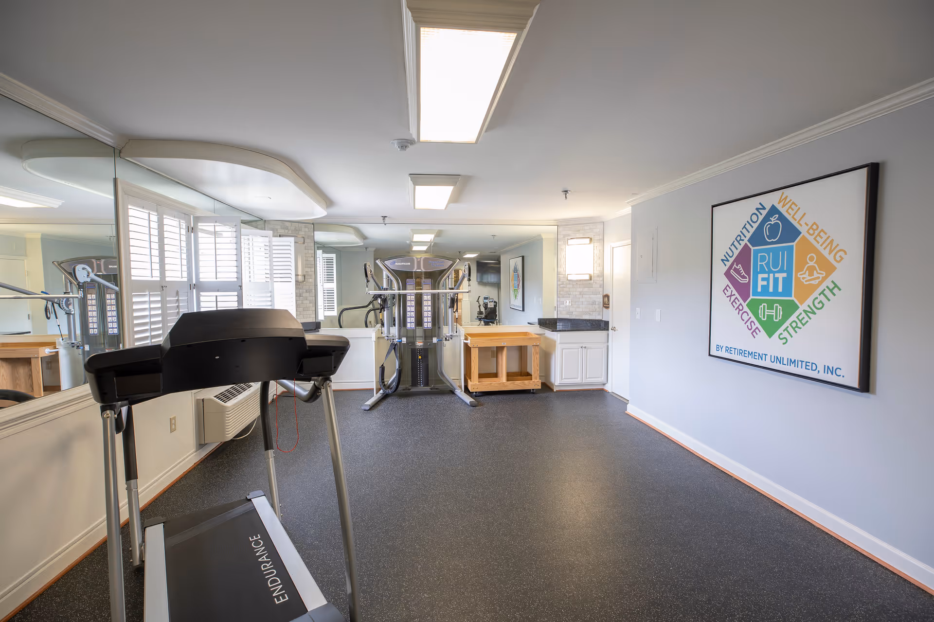 A fitness room with exercise equipment including a treadmill and a multi-functional workout machine. The room has large mirrors on one wall, windows with white shutters, and a framed poster on the opposite wall promoting nutrition, well-being, exercise, and strength. The floor is covered with black rubber matting.