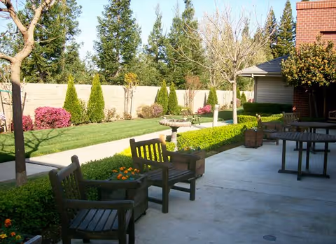 Outdoor patio area with wooden benches and tables, surrounded by manicured bushes, trees, and a lawn with a stone birdbath. A brick building is partially visible on the right side, and a tall fence with greenery is in the background.