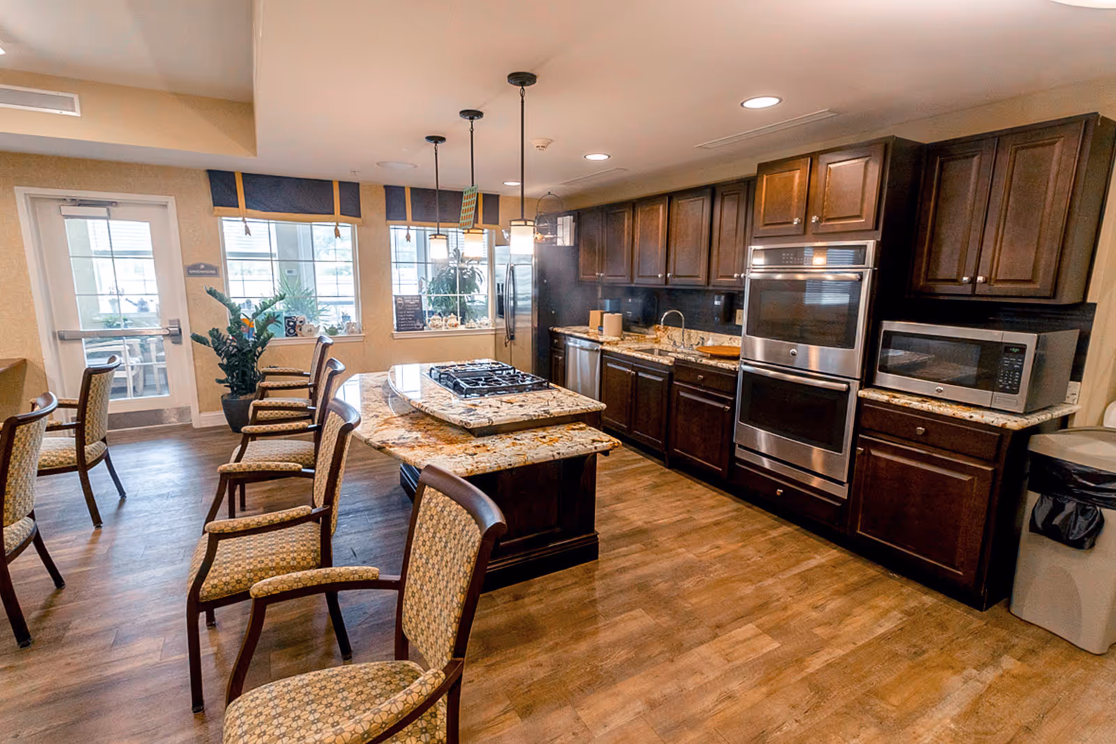 A spacious kitchen area with dark wooden cabinets, granite countertops, and stainless steel appliances including a double oven, microwave, and refrigerator. There is a kitchen island with a built-in gas stove and several upholstered chairs arranged around it. Large windows let in natural light, and there are pendant lights hanging above the island. The floor is wood, and there is a door leading outside.