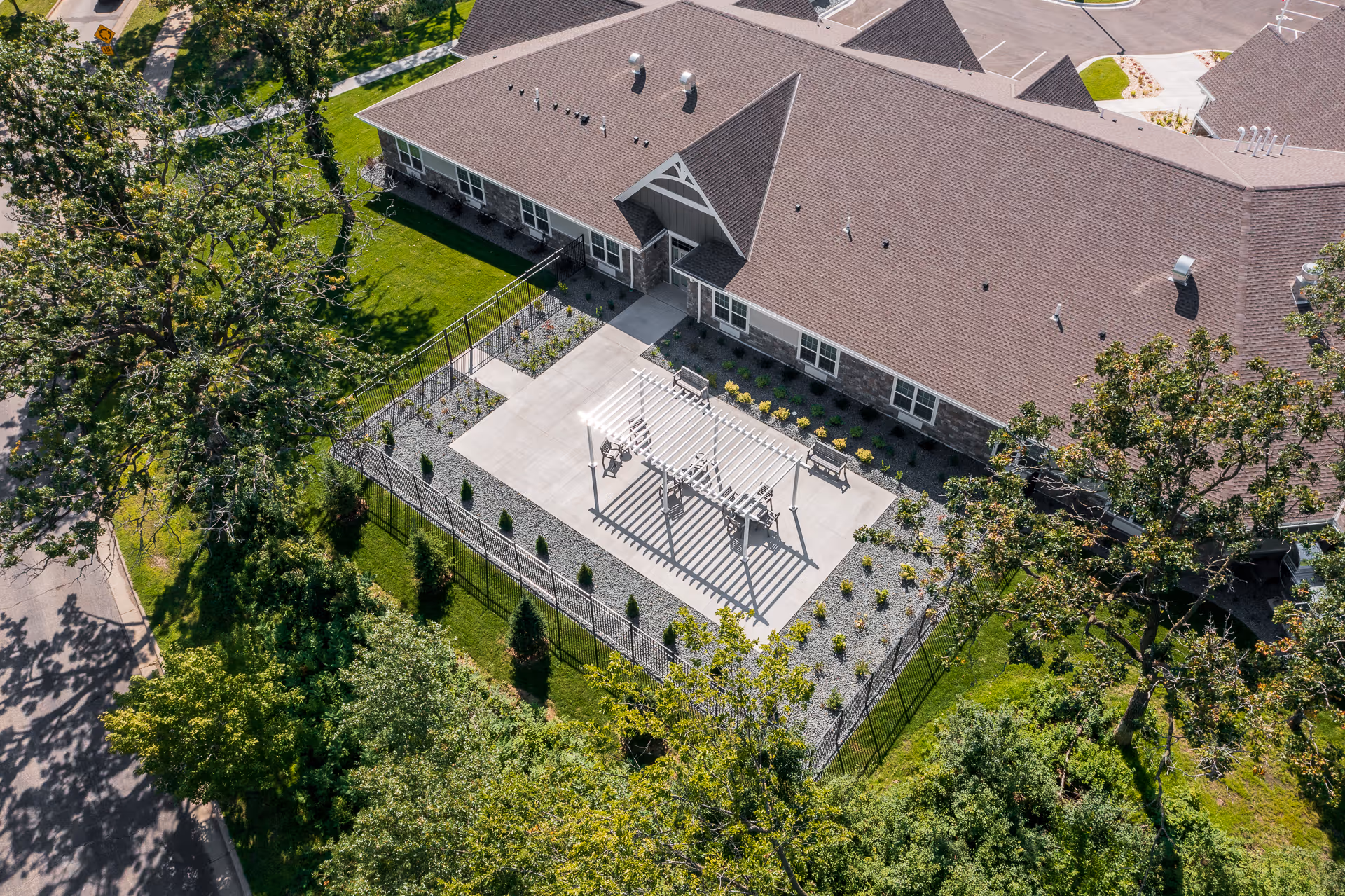 Aerial view of a senior living facility building with a fenced outdoor patio area featuring a pergola, benches, and landscaped greenery surrounding the building.
