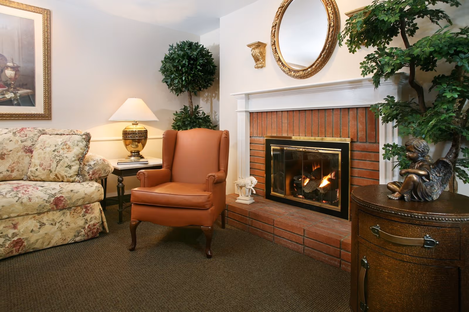 Cozy living room area with a floral patterned sofa, a brown leather armchair, a side table with a gold lamp, a brick fireplace with a fire burning, a round gold-framed mirror above the fireplace, and decorative plants and statues.