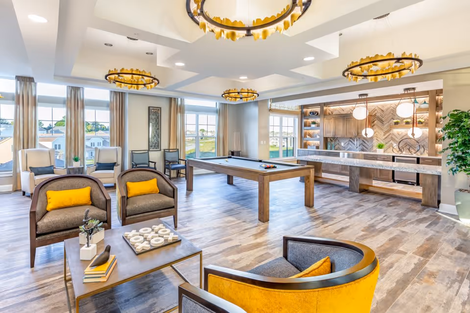 A bright and spacious senior living common area featuring a pool table in the center, surrounded by comfortable seating including armchairs with yellow cushions and a coffee table with books and decorative items. Large windows allow natural light to fill the room, and a modern kitchenette with pendant lights and wooden cabinetry is visible in the background. The room has stylish circular chandeliers and wood-patterned flooring.