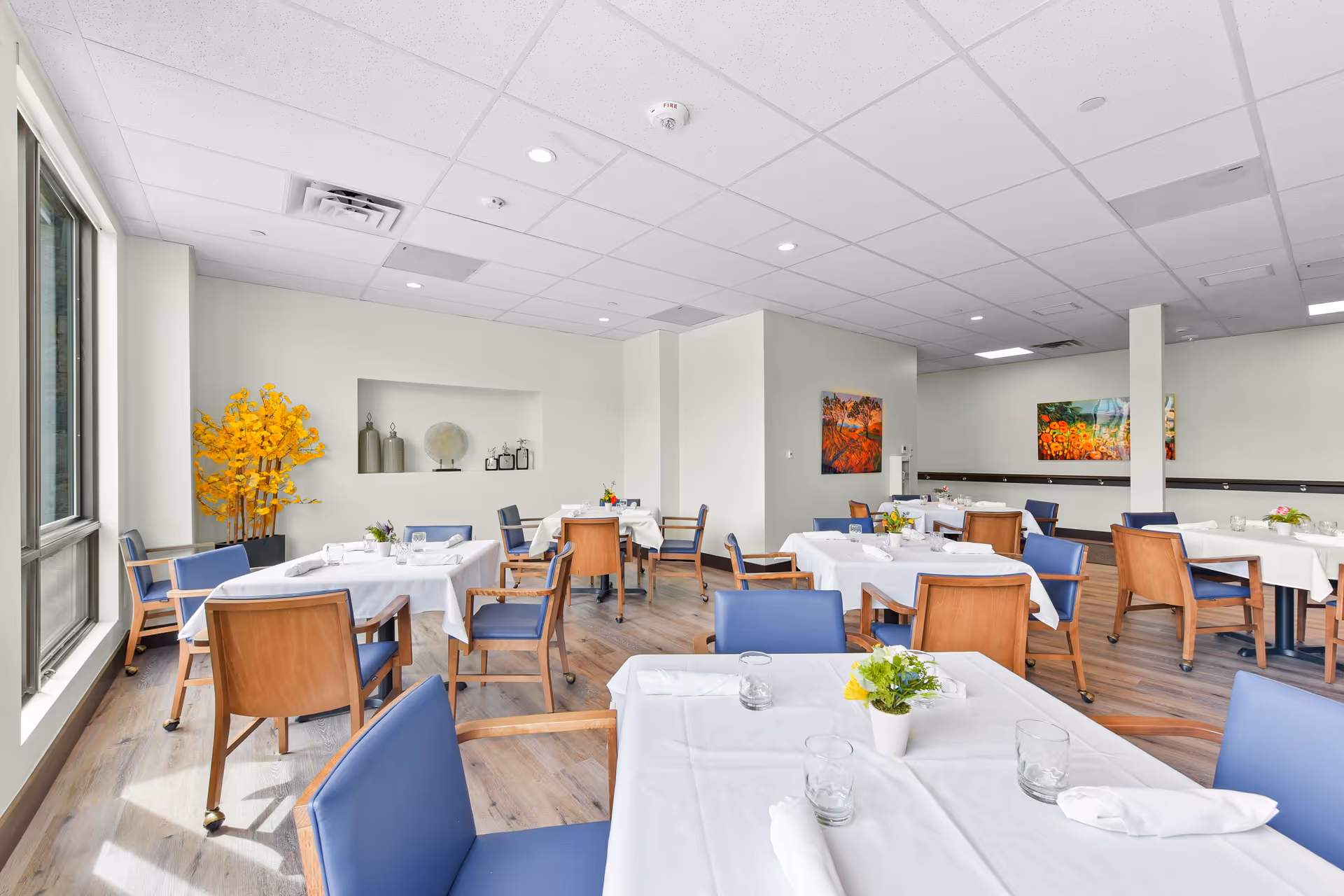 Bright senior living dining room with tables covered in white tablecloths, blue chairs, and small floral centerpieces.