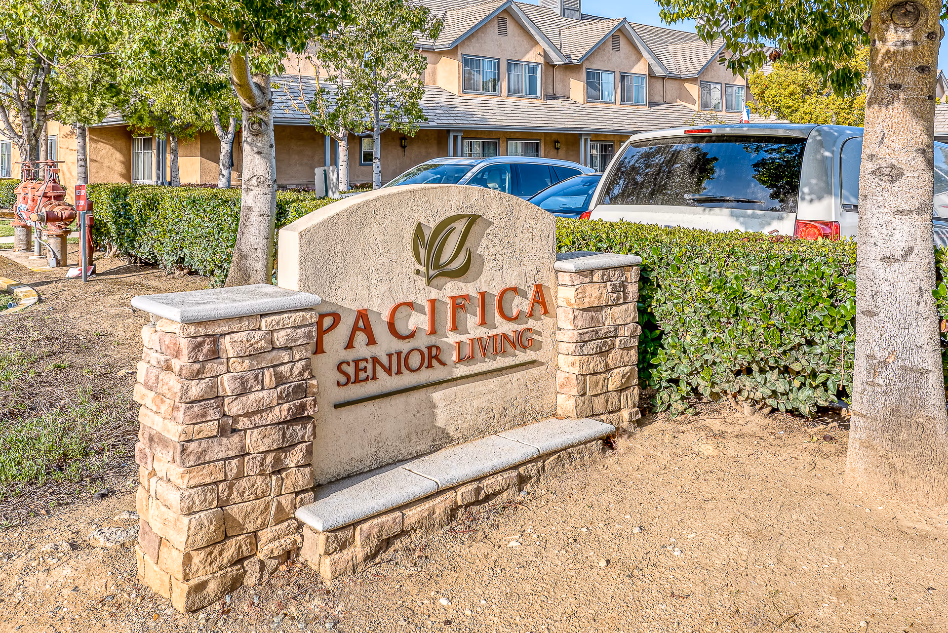 Outdoor view of the entrance sign for Pacifica Senior Living, surrounded by trees, bushes, and parked cars, with a residential-style building in the background.