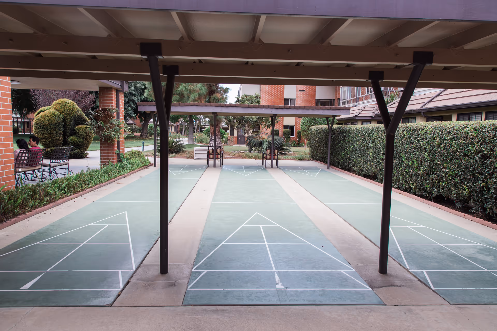 Covered outdoor shuffleboard courts with green playing surfaces and white markings, surrounded by hedges and brick buildings. Two people are sitting on a bench to the left side of the image.