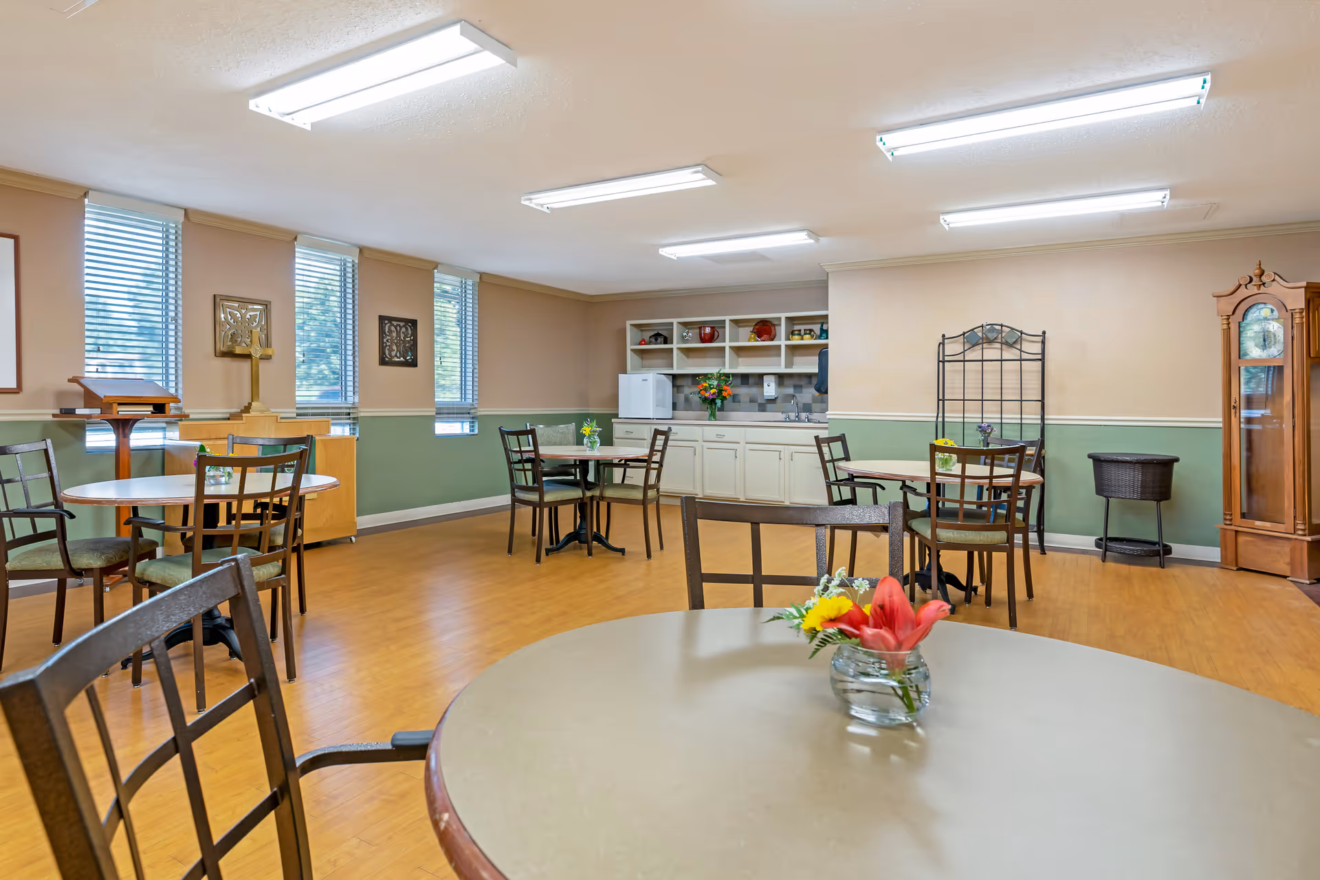 A well-lit dining room in a senior living facility with several round tables and chairs. Each table has a small flower arrangement. The room features large windows with blinds, a kitchenette area with cabinets, a refrigerator, and a sink. There is also a wooden grandfather clock and decorative wall art.